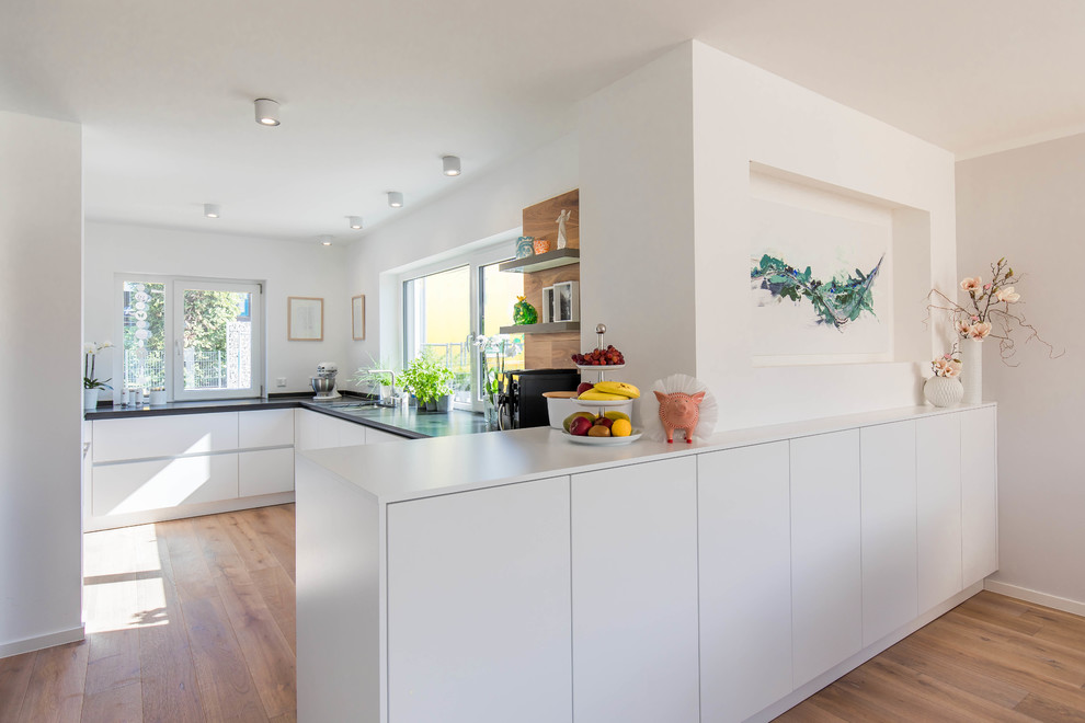 This is an example of a mid-sized scandinavian u-shaped open plan kitchen in Nuremberg with a drop-in sink, flat-panel cabinets, white cabinets, medium hardwood floors, a peninsula, brown floor and black benchtop.