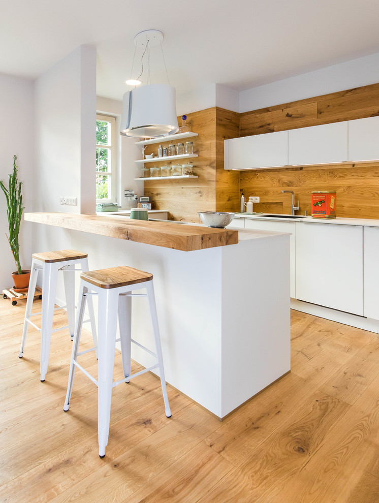 Bild på ett mellanstort skandinaviskt vit vitt kök, med en integrerad diskho, släta luckor, vita skåp, brunt stänkskydd, stänkskydd i trä, mellanmörkt trägolv, en halv köksö och brunt golv
