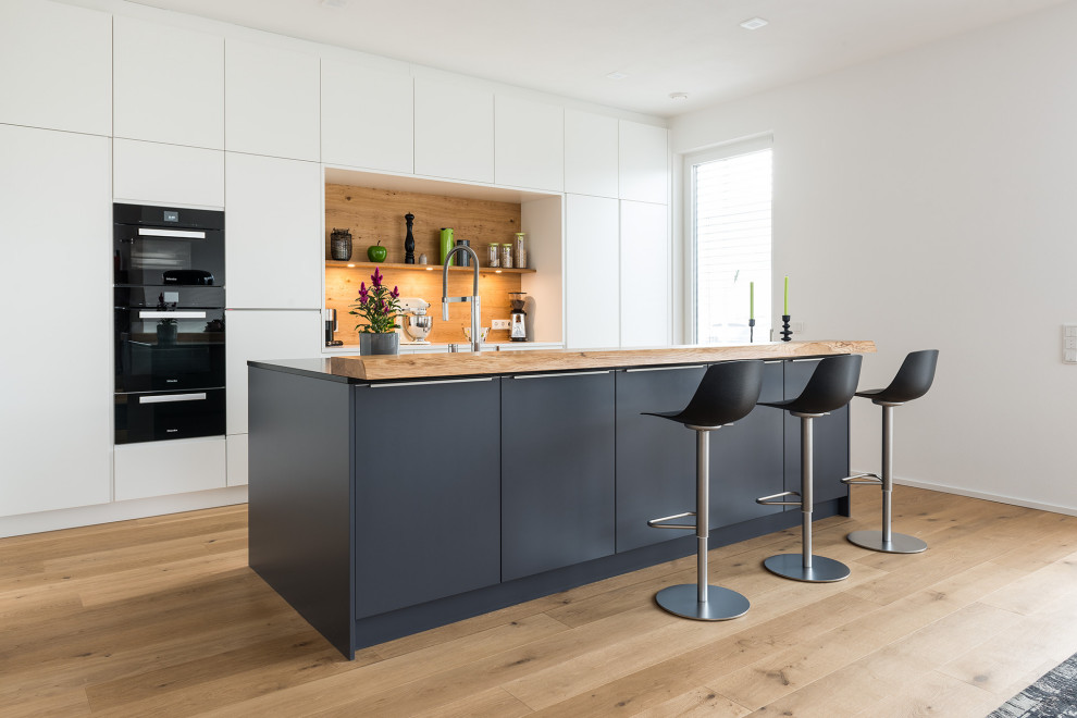 This is an example of a large contemporary kitchen/diner in Munich with a submerged sink, flat-panel cabinets, white cabinets, granite worktops, brown splashback, wood splashback, black appliances, medium hardwood flooring, an island, brown floors and black worktops.