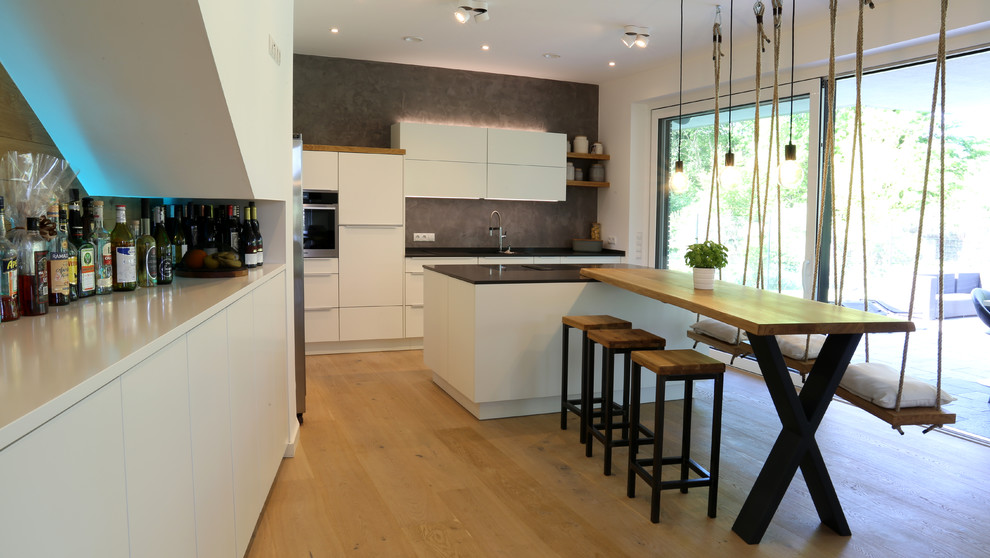 This is an example of a large contemporary single-wall open plan kitchen in Bremen with flat-panel cabinets, white cabinets, grey splashback, stainless steel appliances, medium hardwood flooring, an island, brown floors, black worktops, an integrated sink, granite worktops and stone slab splashback.
