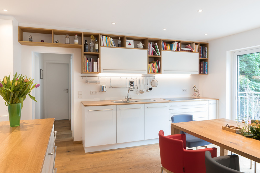 Mid-sized trendy l-shaped medium tone wood floor and brown floor eat-in kitchen photo in Stuttgart with a double-bowl sink, flat-panel cabinets, white cabinets, wood countertops, white backsplash, glass sheet backsplash, stainless steel appliances, brown countertops and an island