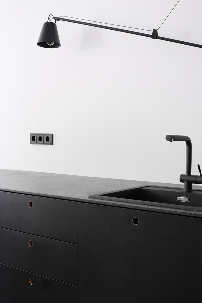Ejemplo de cocina lineal actual pequeña abierta con armarios con paneles lisos, puertas de armario negras, encimera de acrílico, electrodomésticos negros y suelo de madera clara