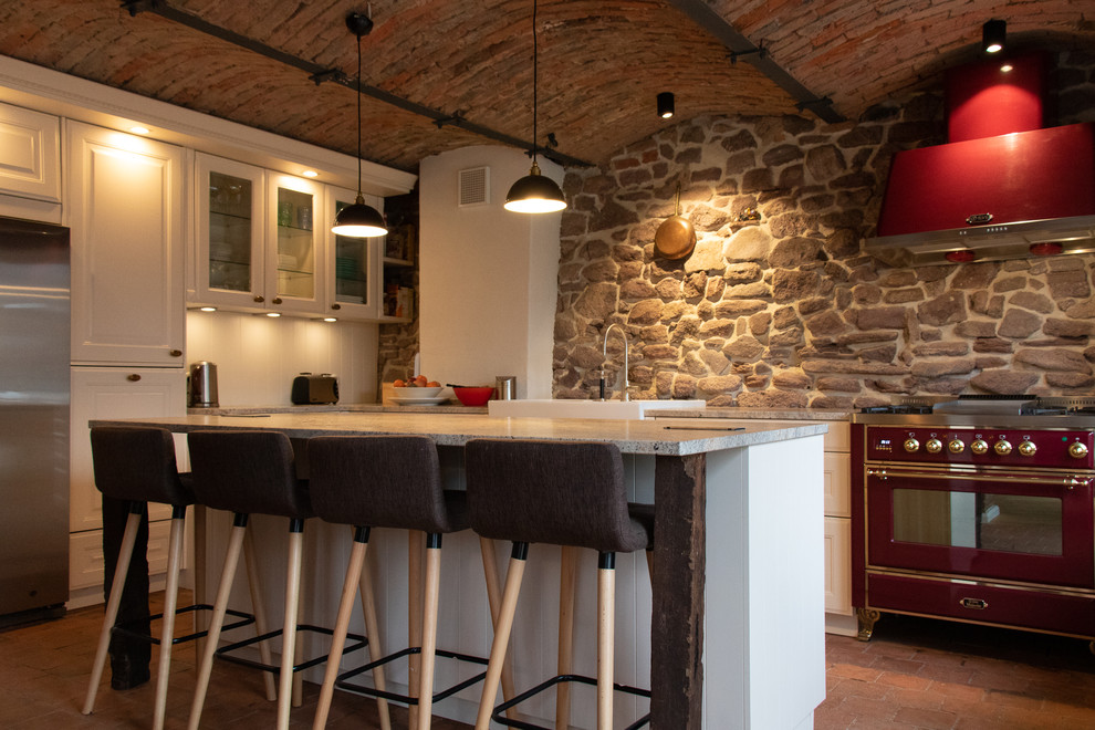 Immagine di un cucina con isola centrale mediterraneo con ante con riquadro incassato, ante bianche, paraspruzzi grigio, elettrodomestici colorati, pavimento in mattoni, pavimento rosso e top grigio