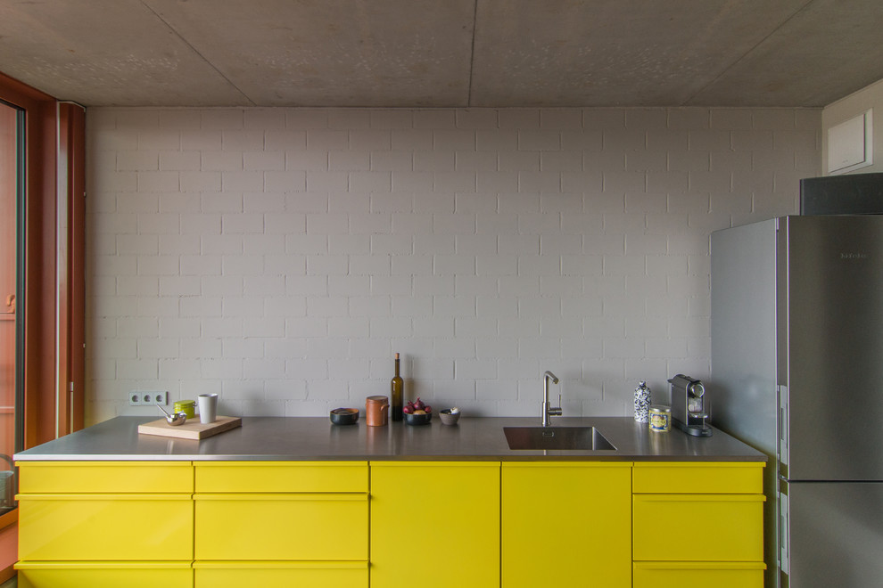 Einzeilige Moderne Wohnküche mit Waschbecken, flächenbündigen Schrankfronten, gelben Schränken, Edelstahl-Arbeitsplatte, Küchenrückwand in Weiß, Küchengeräten aus Edelstahl, Betonboden und Kücheninsel in Berlin
