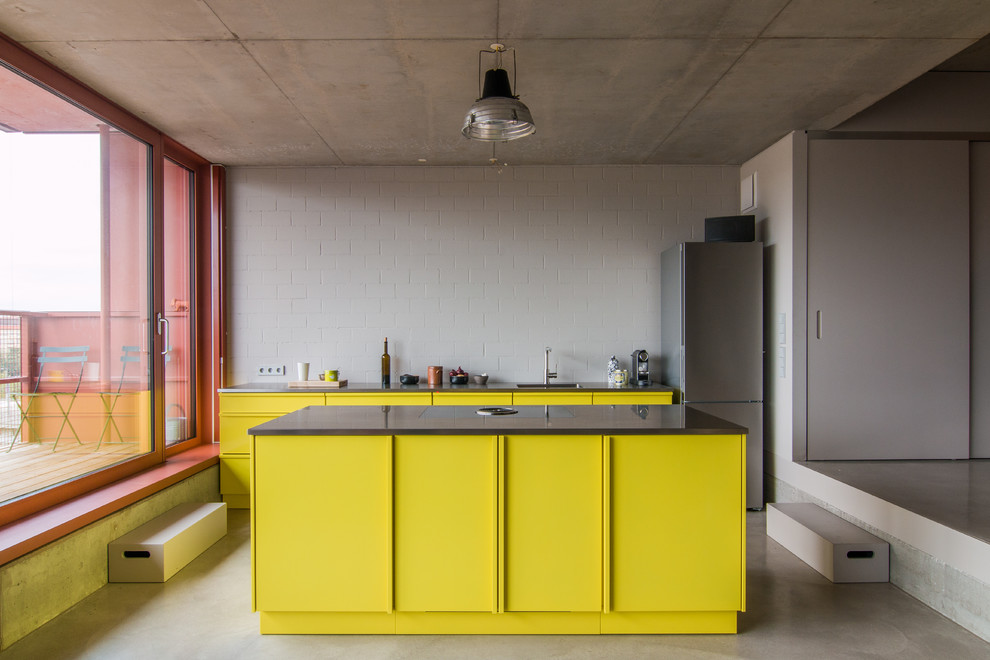 На фото: кухня среднего размера в стиле лофт с одинарной мойкой, плоскими фасадами, желтыми фасадами, техникой из нержавеющей стали, бетонным полом, островом и серым фартуком с