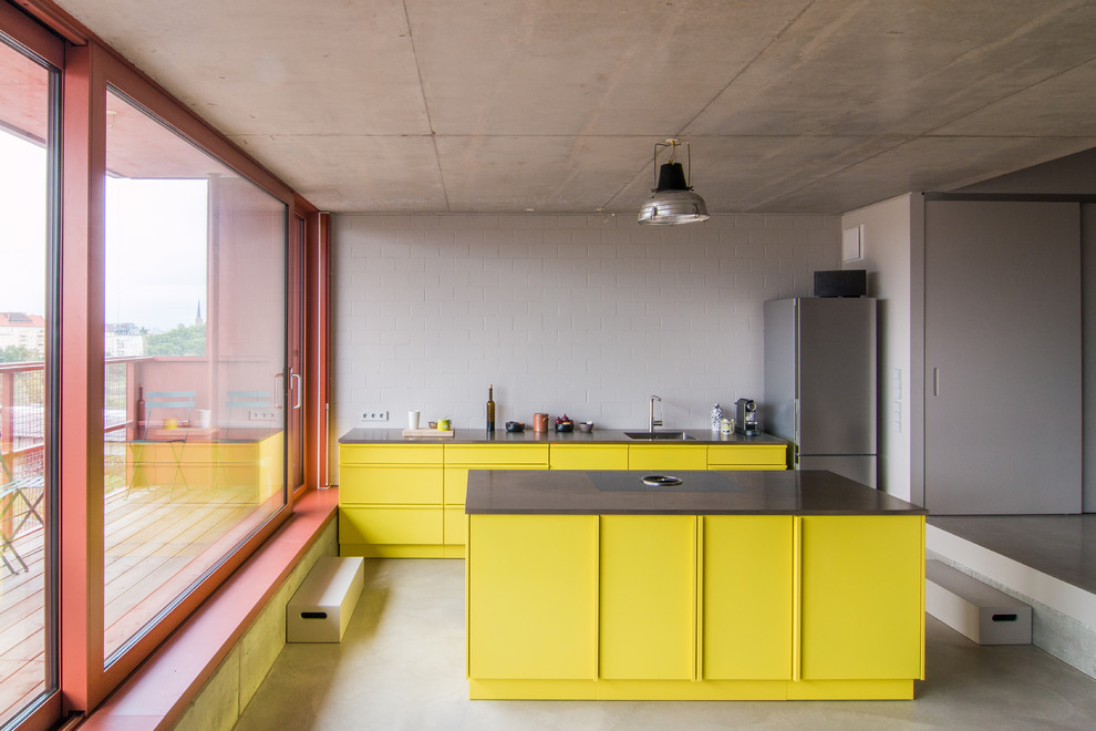 На фото: прямая кухня в современном стиле с обеденным столом, одинарной мойкой, плоскими фасадами, желтыми фасадами, столешницей из нержавеющей стали, белым фартуком, техникой из нержавеющей стали, бетонным полом и островом