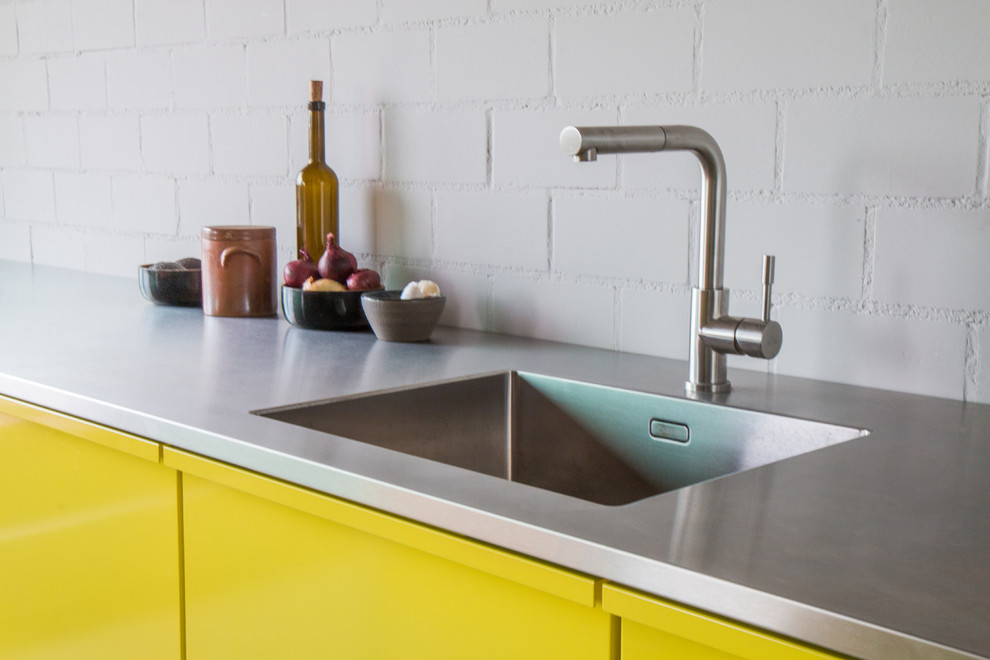 Idee per una cucina design con lavello a vasca singola, ante lisce, ante gialle, top in acciaio inossidabile, paraspruzzi bianco, elettrodomestici in acciaio inossidabile e pavimento in cemento