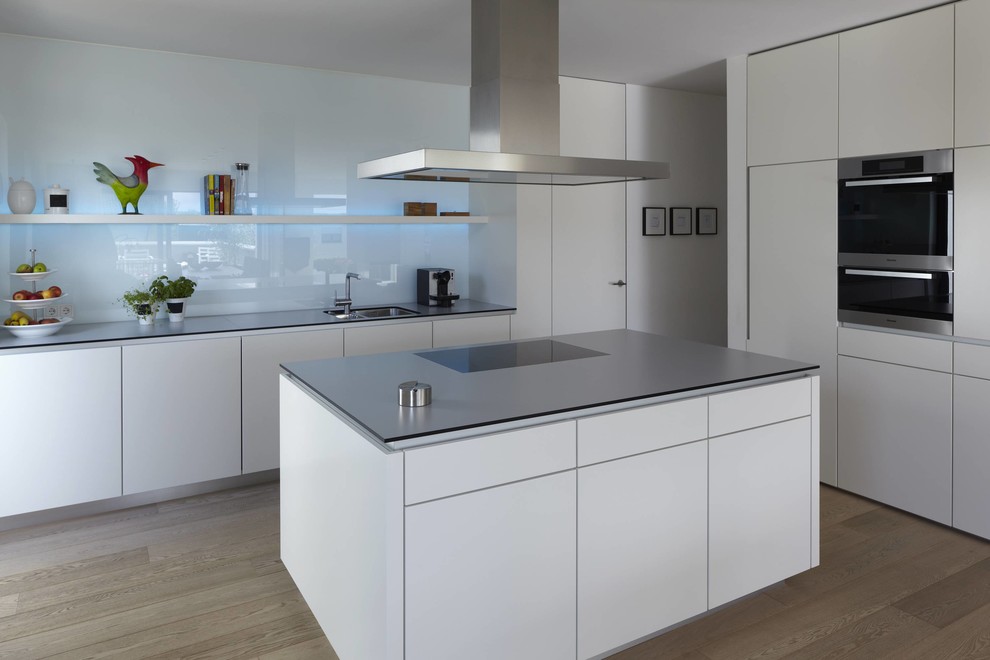 This is an example of a large contemporary kitchen in Other with a submerged sink, flat-panel cabinets, white cabinets, white splashback, glass sheet splashback, stainless steel appliances, medium hardwood flooring and an island.