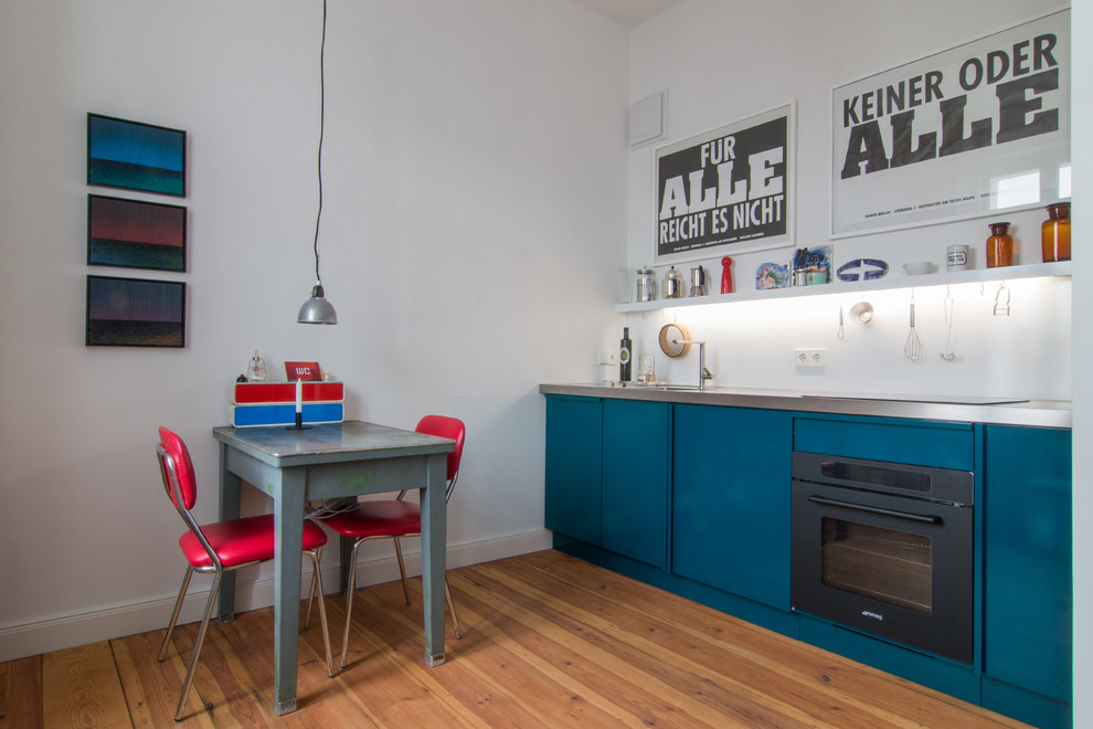 Industriell inredning av ett litet linjärt kök och matrum, med släta luckor, blå skåp, bänkskiva i rostfritt stål, vitt stänkskydd, svarta vitvaror, mellanmörkt trägolv och brunt golv