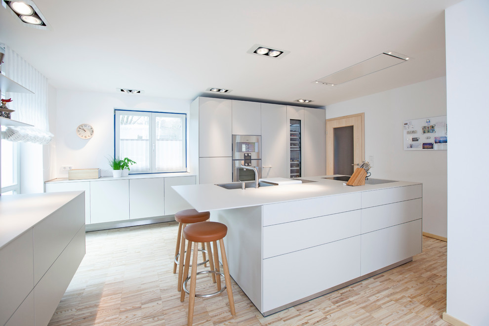 Photo of a large contemporary galley open plan kitchen in Nuremberg with a built-in sink, flat-panel cabinets, white cabinets, white splashback, stainless steel appliances, light hardwood flooring and an island.