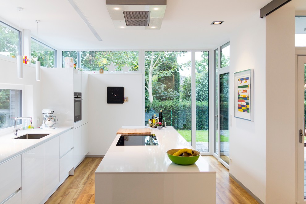 Einzeilige Moderne Küche mit Einbauwaschbecken, flächenbündigen Schrankfronten, weißen Schränken, Küchenrückwand in Weiß, schwarzen Elektrogeräten, braunem Holzboden und Kücheninsel in Sonstige