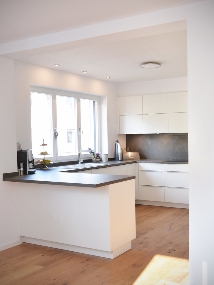 Photo of an expansive contemporary u-shaped open plan kitchen in Frankfurt with flat-panel cabinets, white cabinets, grey splashback, brown floors, grey worktops, a built-in sink, wood worktops, wood splashback, stainless steel appliances, light hardwood flooring and no island.