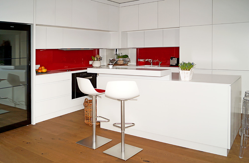 Идея дизайна: угловая кухня-гостиная среднего размера в современном стиле с двойной мойкой, плоскими фасадами, белыми фасадами, столешницей из акрилового камня, красным фартуком, фартуком из стекла, черной техникой, деревянным полом и островом
