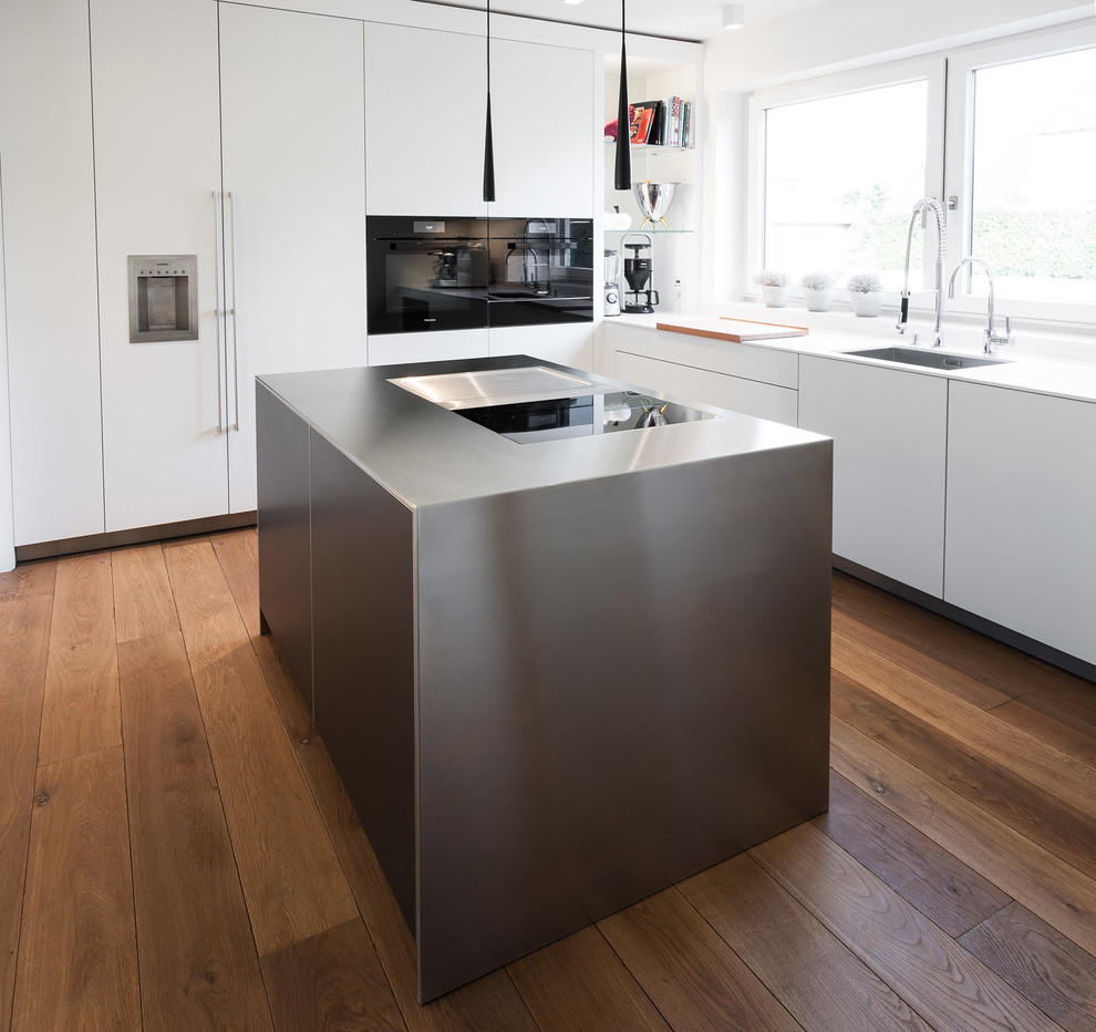 Photo of a medium sized contemporary l-shaped enclosed kitchen in Dusseldorf with an integrated sink, flat-panel cabinets, white cabinets, black appliances, dark hardwood flooring, an island, brown floors and white worktops.