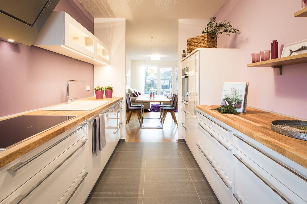 Пример оригинального дизайна: отдельная, параллельная, глянцевая кухня среднего размера в современном стиле с накладной мойкой, плоскими фасадами, белыми фасадами, деревянной столешницей, техникой под мебельный фасад и розовым фартуком без острова