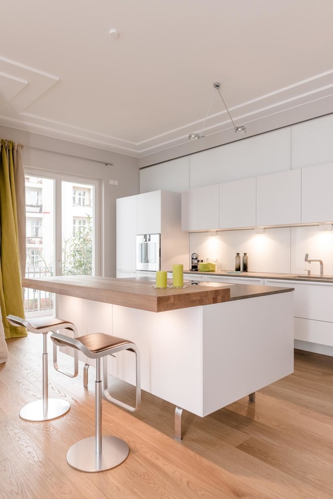 Inspiration for a large contemporary single-wall open plan kitchen in Berlin with flat-panel cabinets, white cabinets, quartz worktops, white splashback, glass sheet splashback, stainless steel appliances, medium hardwood flooring, an island, beige floors, brown worktops and a submerged sink.