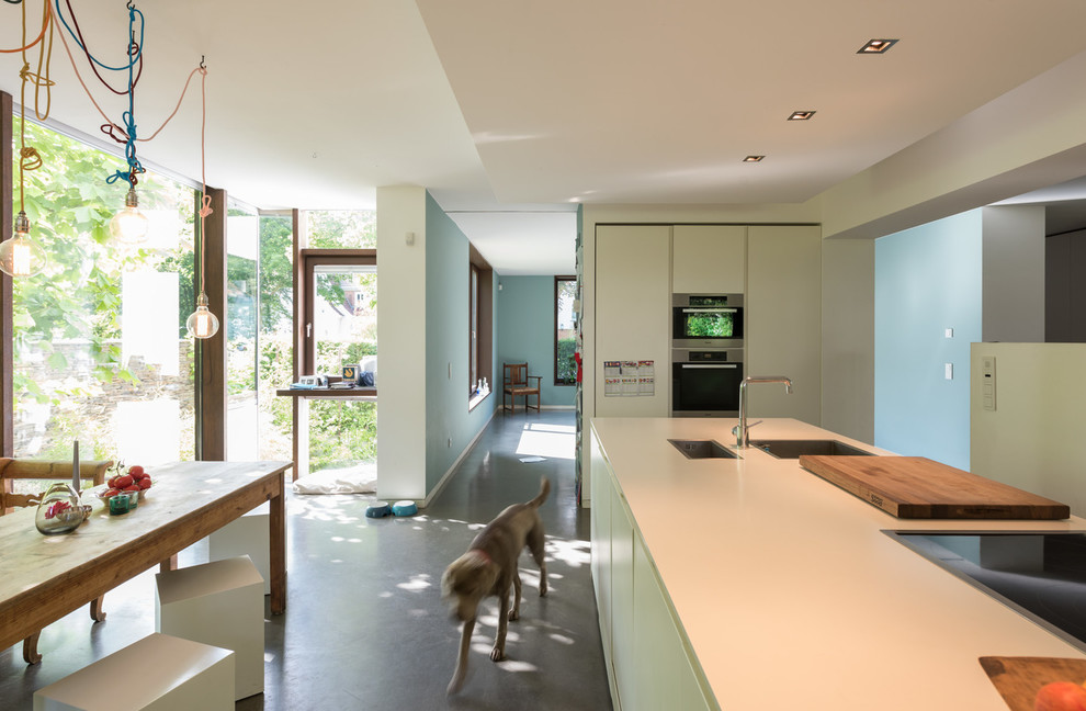 Идея дизайна: прямая кухня-гостиная среднего размера в современном стиле с накладной мойкой, плоскими фасадами, бирюзовыми фасадами, черной техникой, бетонным полом и серым полом