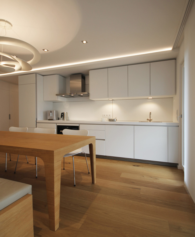 Design ideas for a medium sized contemporary single-wall open plan kitchen in Munich with a built-in sink, flat-panel cabinets, white cabinets, laminate countertops, white splashback, glass sheet splashback, stainless steel appliances, light hardwood flooring, brown floors and white worktops.