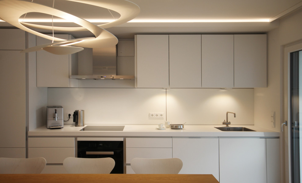 Photo of a medium sized contemporary single-wall open plan kitchen in Munich with a built-in sink, flat-panel cabinets, white cabinets, laminate countertops, white splashback, glass sheet splashback, stainless steel appliances, light hardwood flooring, brown floors and white worktops.