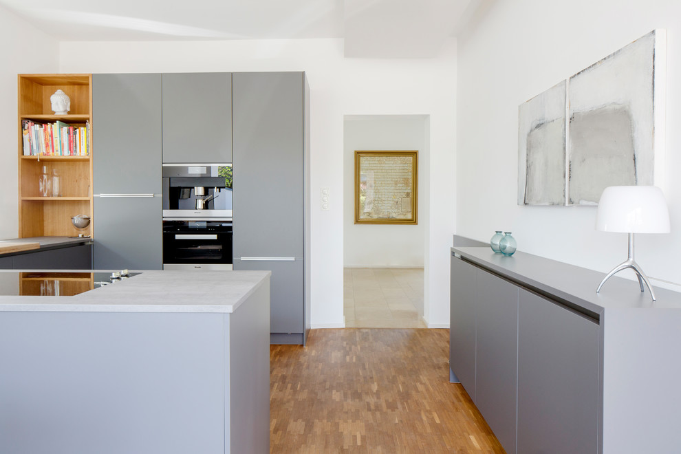 Offene Moderne Küche mit grauen Schränken, Küchenrückwand in Grau, Kücheninsel, braunem Boden, grauer Arbeitsplatte, schwarzen Elektrogeräten und braunem Holzboden in Düsseldorf