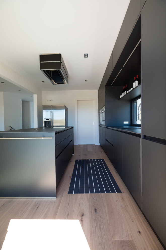 Exemple d'une cuisine ouverte parallèle moderne de taille moyenne avec des portes de placard noires, îlot, un placard à porte plane, fenêtre, un électroménager en acier inoxydable, parquet clair, un sol beige et plan de travail noir.