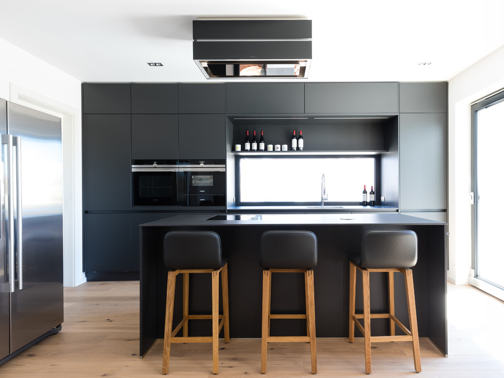 Exempel på ett mellanstort modernt svart linjärt svart kök med öppen planlösning, med en enkel diskho, svarta skåp, svarta vitvaror, en köksö, släta luckor, fönster som stänkskydd, ljust trägolv och brunt golv