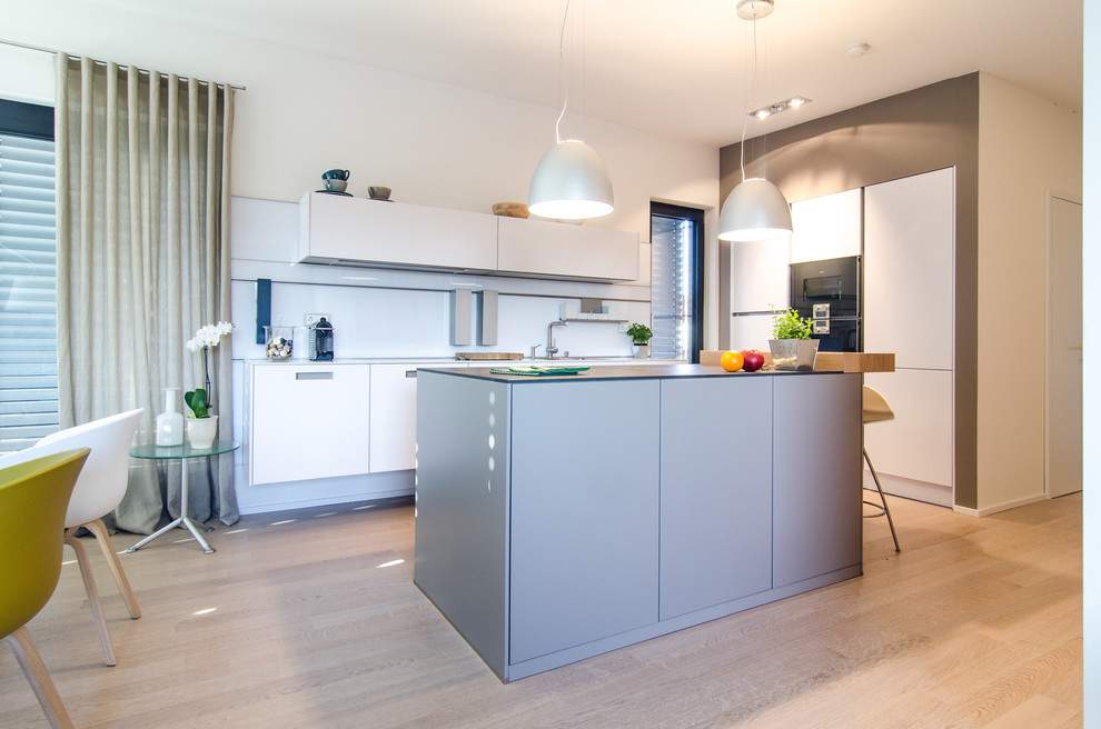 Foto de cocina contemporánea pequeña con una isla, armarios con paneles lisos, salpicadero blanco, electrodomésticos negros, suelo de madera clara y suelo beige
