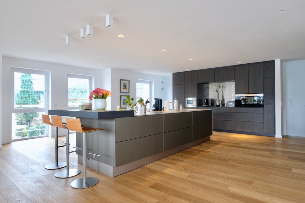 Cette image montre une grande cuisine ouverte linéaire design en bois foncé avec îlot, un placard à porte plane, une crédence métallisée, un électroménager noir, un sol en bois brun et un sol marron.
