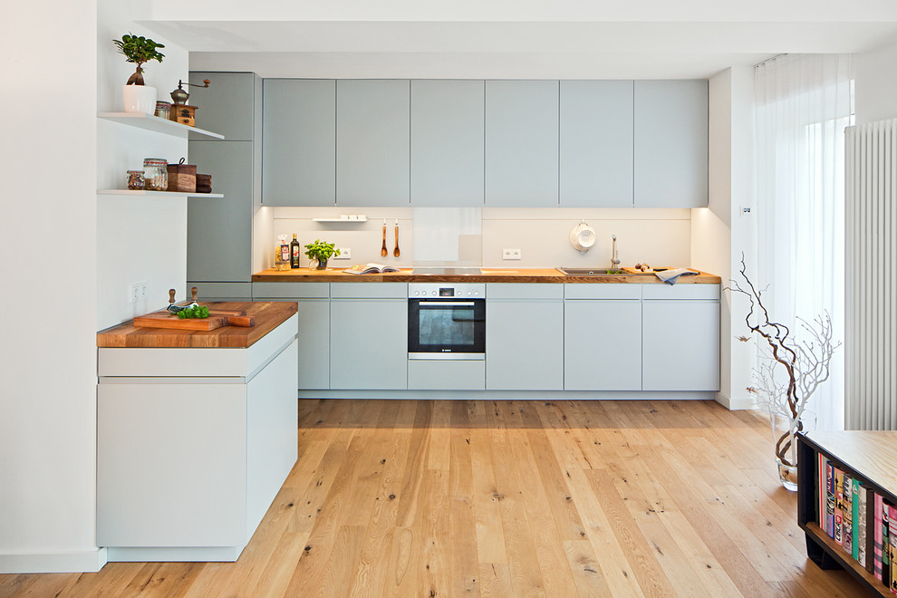 Geschlossene Moderne Küche mit flächenbündigen Schrankfronten, blauen Schränken, hellem Holzboden, Arbeitsplatte aus Holz, Einbauwaschbecken, Küchenrückwand in Weiß, Glasrückwand und Küchengeräten aus Edelstahl in Düsseldorf