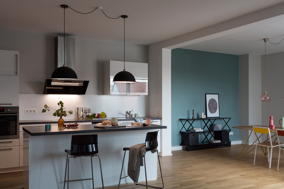Inspiration for an expansive contemporary galley open plan kitchen in Berlin with medium hardwood flooring, brown floors, a built-in sink, flat-panel cabinets, white cabinets, white splashback, stainless steel appliances, an island, black worktops, marble worktops and cement tile splashback.