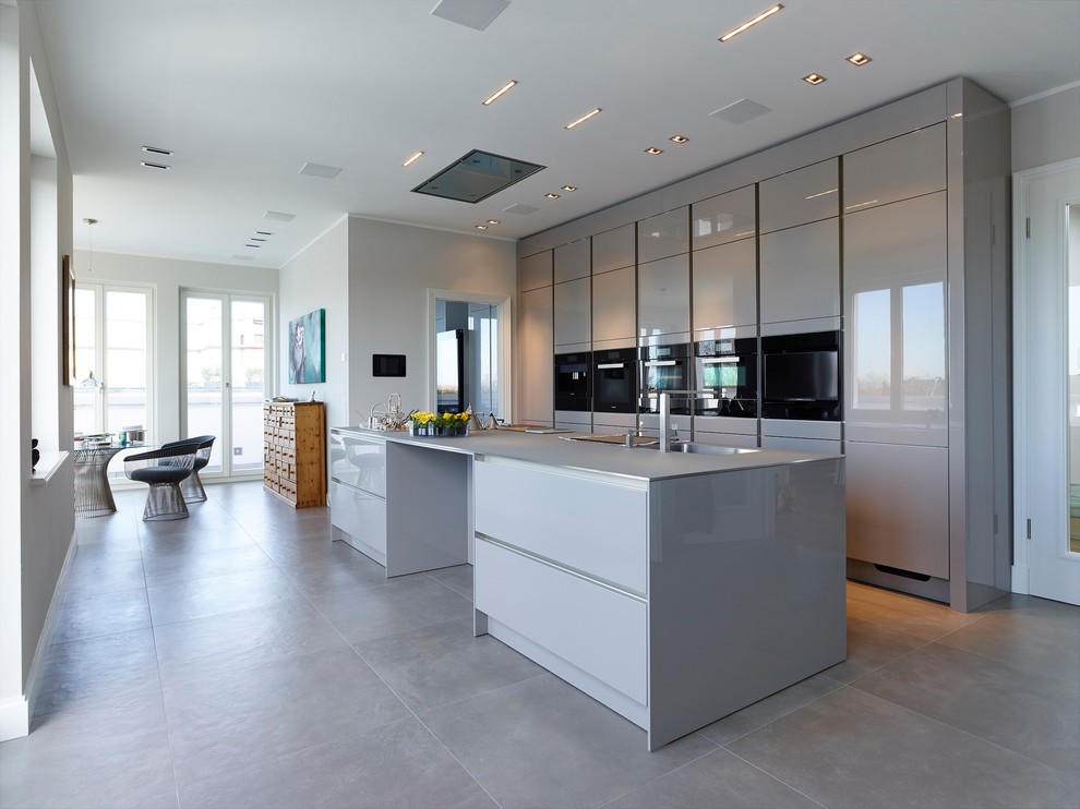 На фото: большая параллельная кухня-гостиная в современном стиле с монолитной мойкой, плоскими фасадами, серыми фасадами, черной техникой, полом из цементной плитки, островом и серым полом