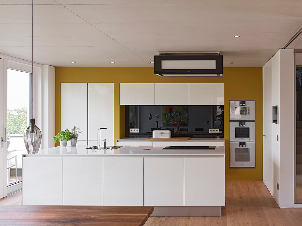 Design ideas for a medium sized contemporary galley open plan kitchen in Stuttgart with a submerged sink, flat-panel cabinets, white cabinets, black splashback, glass sheet splashback, integrated appliances, medium hardwood flooring, an island and brown floors.