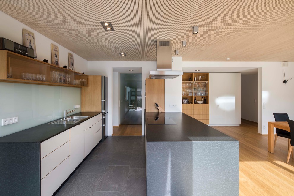 Large minimalist galley black floor and cement tile floor open concept kitchen photo in Munich with white cabinets, an island, a drop-in sink, flat-panel cabinets, blue backsplash and glass sheet backsplash
