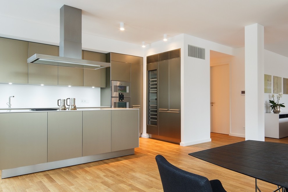 Design ideas for a large contemporary l-shaped open plan kitchen in Berlin with white splashback, light hardwood flooring, flat-panel cabinets, grey cabinets, stainless steel appliances, an island, a built-in sink, glass worktops, glass sheet splashback and beige floors.