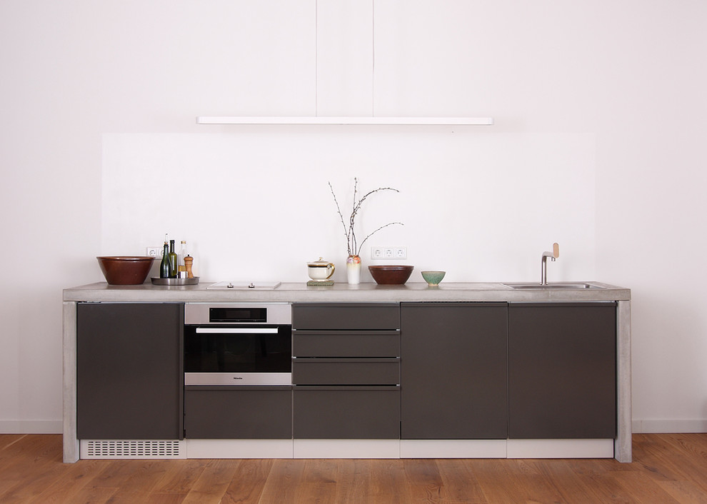 Example of a small trendy single-wall medium tone wood floor and brown floor kitchen design in Berlin with a drop-in sink, flat-panel cabinets, gray cabinets, concrete countertops, white backsplash, paneled appliances and no island
