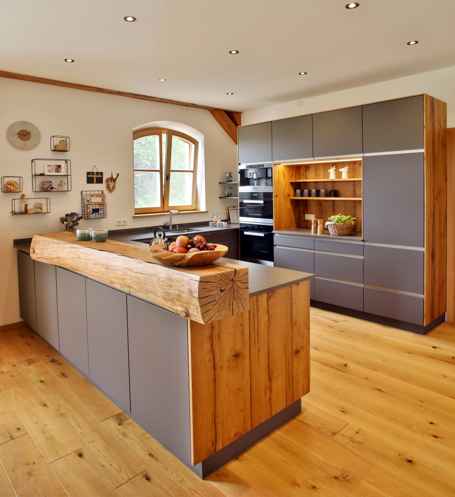 Photo of a farmhouse u-shaped kitchen in Munich with a submerged sink, flat-panel cabinets, grey cabinets, black appliances, medium hardwood flooring, a breakfast bar, brown floors and grey worktops.