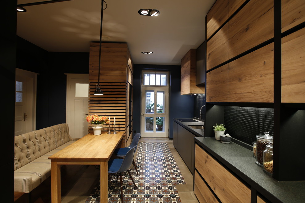 Photo of a medium sized contemporary single-wall kitchen/diner with a built-in sink, flat-panel cabinets, light wood cabinets, granite worktops, black splashback, wood splashback, black appliances, cement flooring, no island and multi-coloured floors.