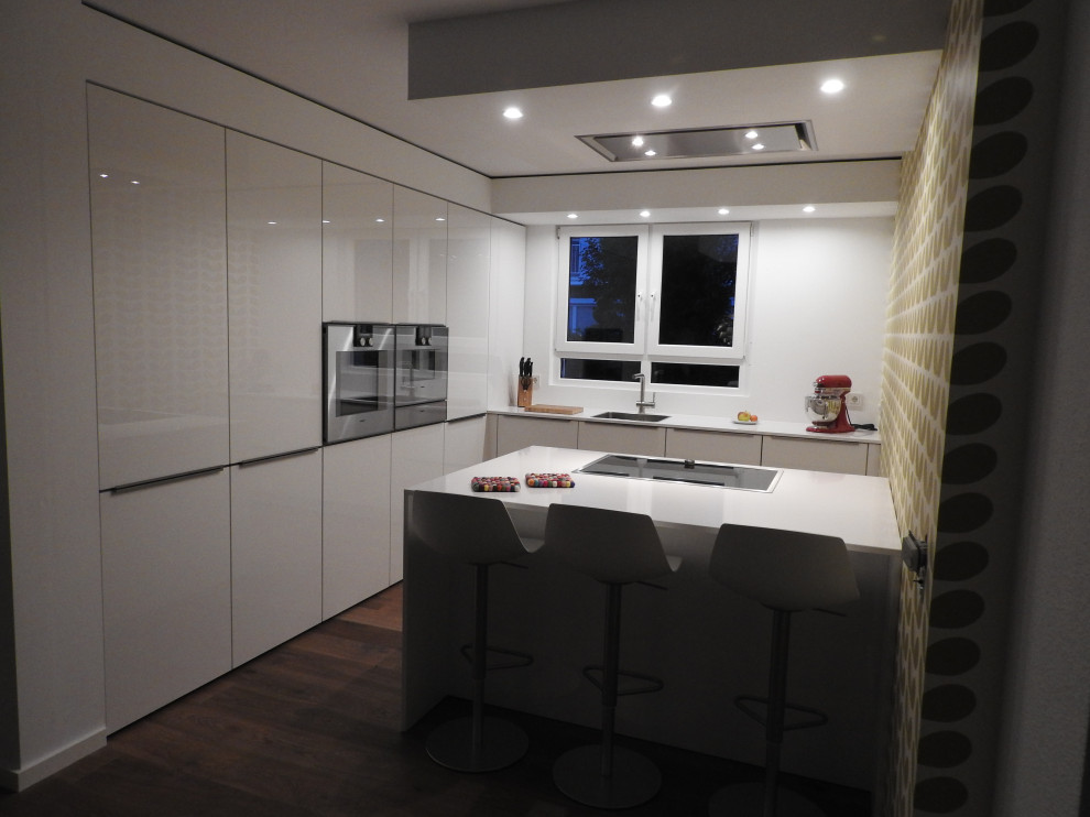 Photo of a medium sized modern l-shaped open plan kitchen in Frankfurt with a built-in sink, flat-panel cabinets, white cabinets, composite countertops, stainless steel appliances, dark hardwood flooring, a breakfast bar, brown floors and white worktops.