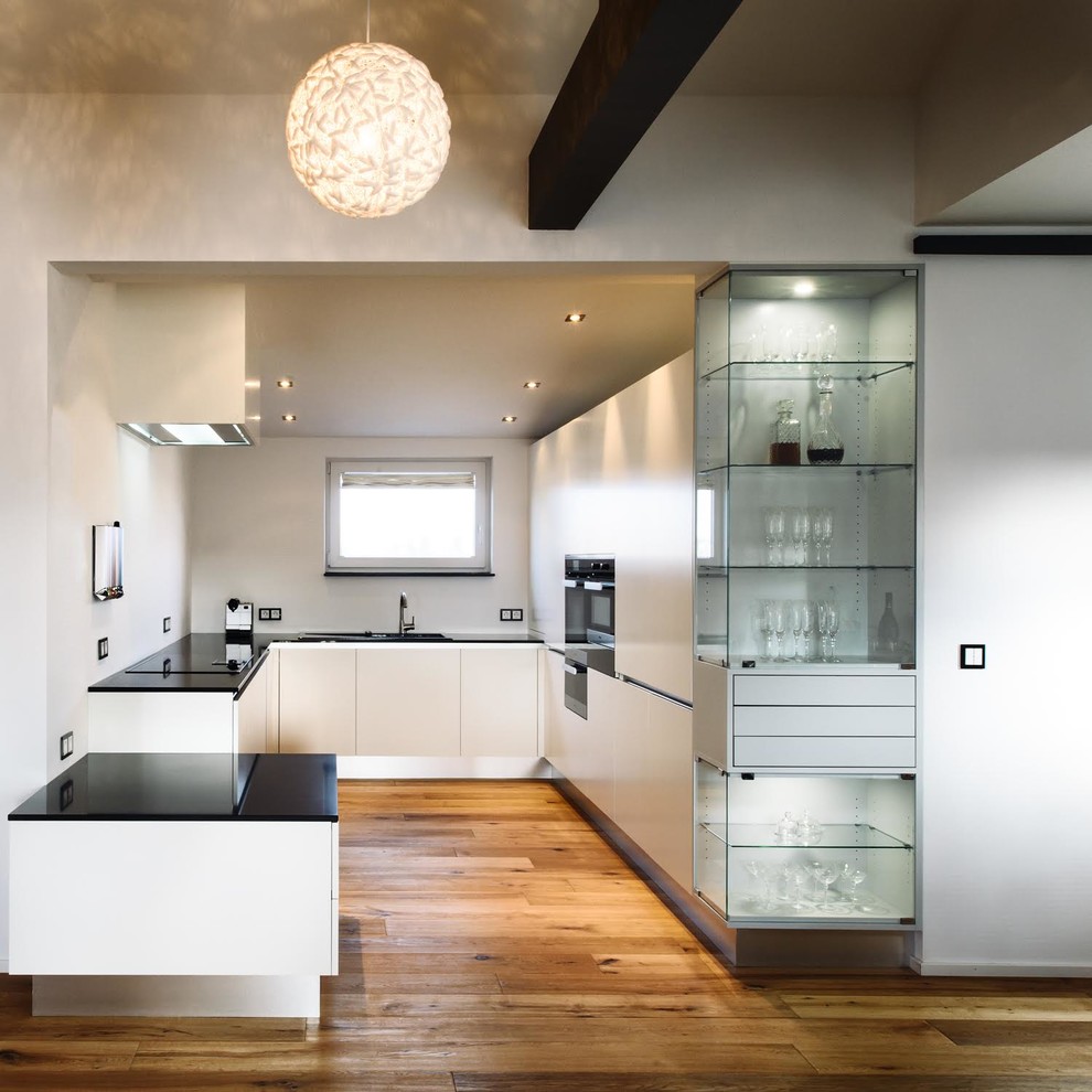 Mittelgroße Moderne Küche in U-Form mit integriertem Waschbecken, flächenbündigen Schrankfronten, weißen Schränken, Küchenrückwand in Weiß, schwarzen Elektrogeräten, braunem Holzboden und Halbinsel in Stuttgart