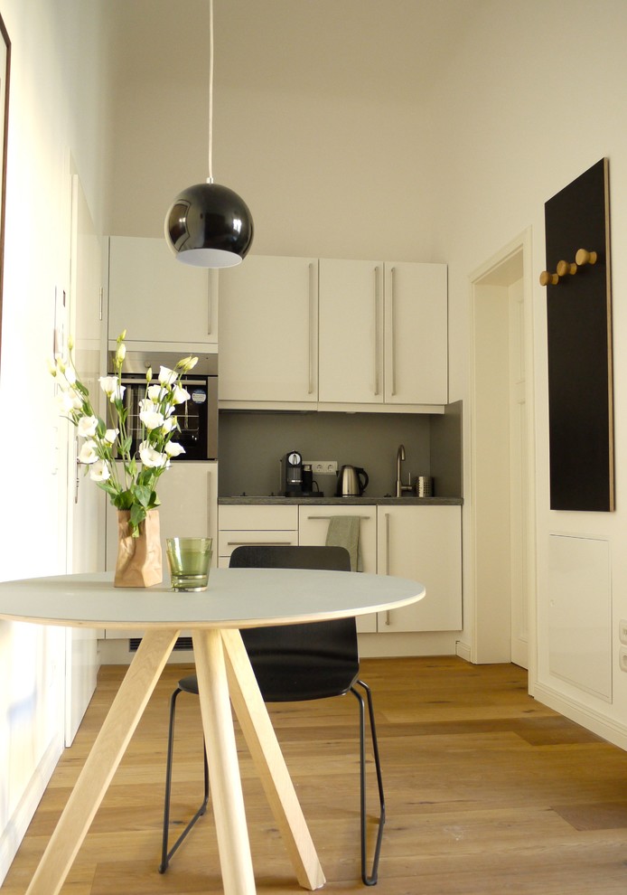 Einzeilige, Kleine Moderne Küche ohne Insel mit flächenbündigen Schrankfronten, weißen Schränken, Küchenrückwand in Grau, Küchengeräten aus Edelstahl und braunem Holzboden in Berlin