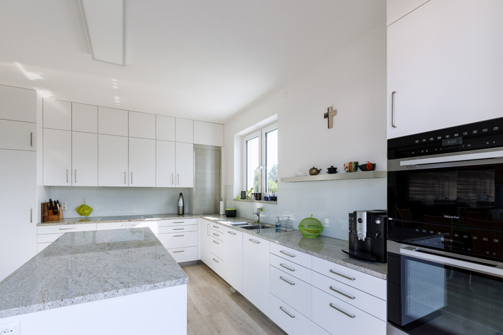 Inspiration for a large contemporary l-shaped open plan kitchen in Stuttgart with a double-bowl sink, flat-panel cabinets, white cabinets, granite worktops, white splashback, glass sheet splashback, black appliances, medium hardwood flooring, an island, beige floors, grey worktops and a wallpapered ceiling.