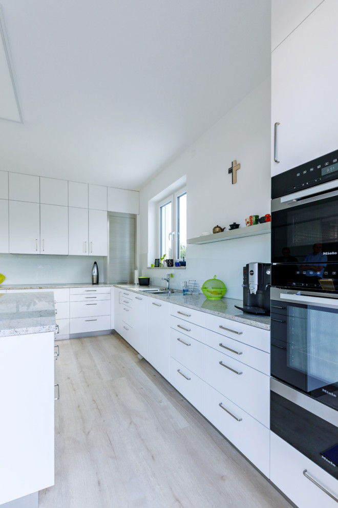 Bild på ett stort funkis grå grått kök, med en dubbel diskho, släta luckor, vita skåp, granitbänkskiva, vitt stänkskydd, glaspanel som stänkskydd, svarta vitvaror, mellanmörkt trägolv, en köksö och beiget golv