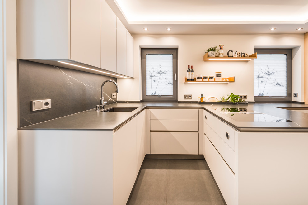 Kleine Moderne Wohnküche in U-Form mit flächenbündigen Schrankfronten, weißen Schränken, Küchenrückwand in Weiß, schwarzen Elektrogeräten, Halbinsel und grauer Arbeitsplatte in Dresden