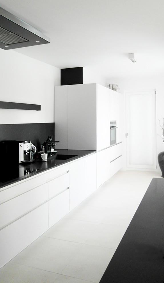 На фото: прямая кухня-гостиная среднего размера с монолитной мойкой, плоскими фасадами, белыми фасадами, черным фартуком и черной техникой без острова