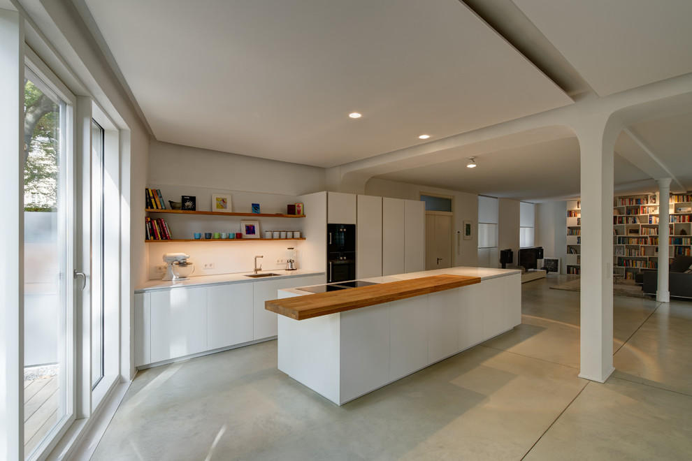 Photo of a medium sized industrial galley open plan kitchen in Frankfurt with a submerged sink, flat-panel cabinets, white cabinets, composite countertops, white splashback, black appliances and an island.
