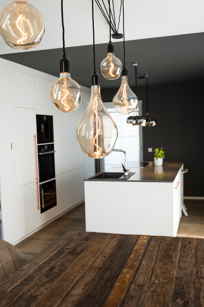 Photo of a modern open plan kitchen in Dusseldorf with flat-panel cabinets, white cabinets, concrete worktops, grey splashback, black appliances, dark hardwood flooring, an island, brown floors and grey worktops.