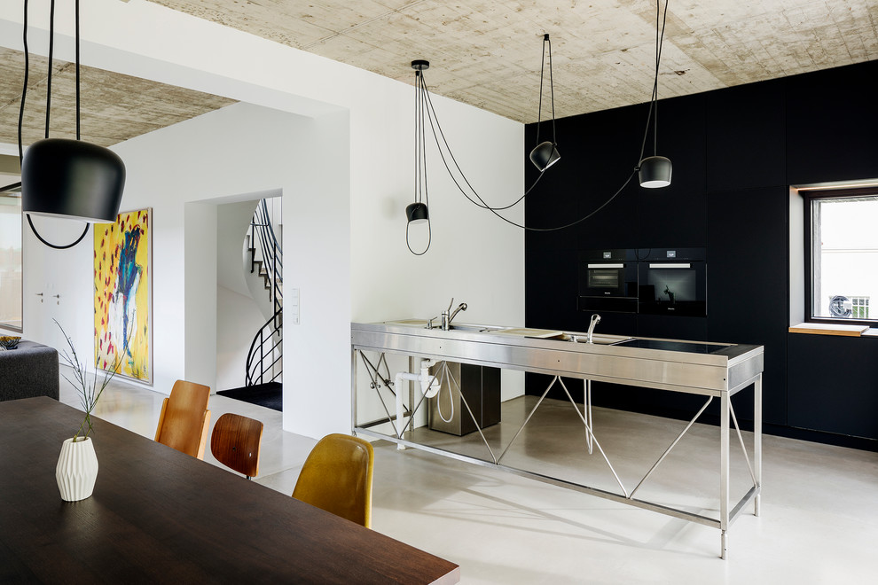 Medium sized industrial galley open plan kitchen in Berlin with an integrated sink, flat-panel cabinets, black cabinets, stainless steel worktops, black appliances, concrete flooring, a breakfast bar, grey floors and grey worktops.