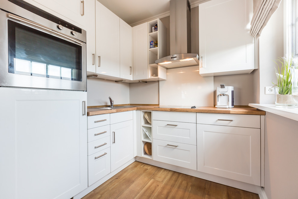 Bild på ett avskilt, litet lantligt l-kök, med en nedsänkt diskho, vita skåp, träbänkskiva, beige stänkskydd, rostfria vitvaror, mellanmörkt trägolv och skåp i shakerstil