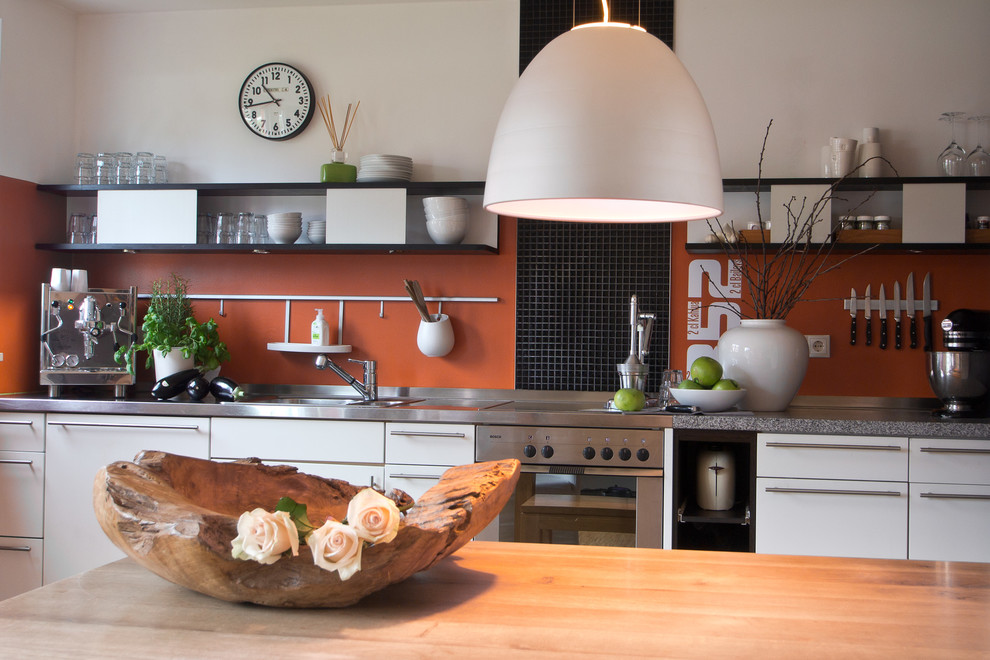 Imagen de cocina lineal actual de tamaño medio abierta con puertas de armario blancas, encimera de acero inoxidable y salpicadero naranja
