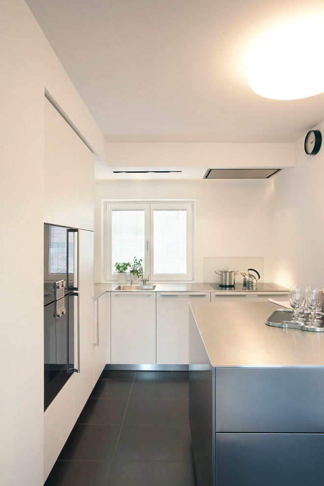 Exempel på ett avskilt, litet modernt kök, med en integrerad diskho, släta luckor, vita skåp, bänkskiva i rostfritt stål, vitt stänkskydd, glaspanel som stänkskydd, svarta vitvaror, en köksö och betonggolv